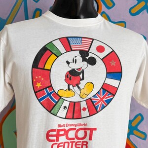 Może przedstawiać: Biała koszulka z Myszka Miki otoczona flagami międzynarodowymi. Tekst "Walt Disney World EPCOT CENTER" jest wydrukowany na czerwono. Koszulka jest prezentowana na czarnym manekinie.