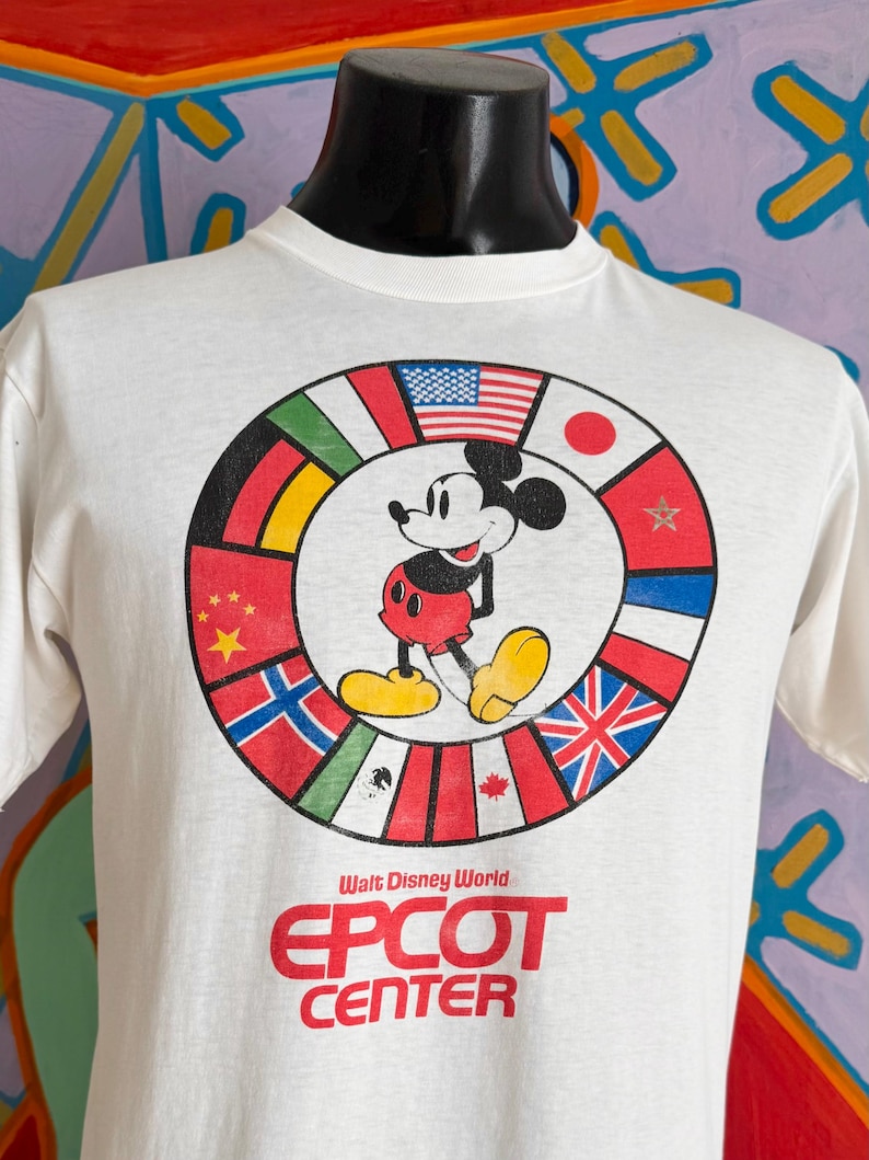Może przedstawiać: Biała koszulka z Myszka Miki otoczona flagami międzynarodowymi. Tekst "Walt Disney World EPCOT CENTER" jest wydrukowany na czerwono. Koszulka jest prezentowana na czarnym manekinie.