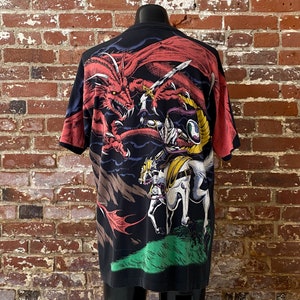 Może przedstawiać: Czarny t-shirt z fantastyczną grafiką przedstawiającą czerwonego smoka, rycerza na białym koniu i wojownika z mieczem. Projekt wykorzystuje żywe kolory na ciemnym tle. T-shirt jest prezentowany na manekinie.