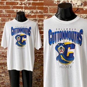 May include: White t-shirt with a blue and yellow graphic of a snake with the text "Columbus Cottonmouths Hockey" printed on the front.