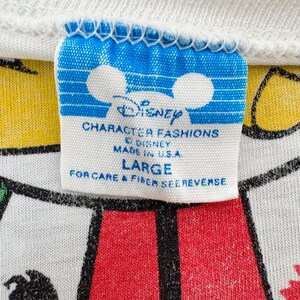Może przedstawiać: Zbliżenie białego kołnierzyka koszulki z niebiesko-białą metką Disney Character Fashions. Na metce widnieje napis "LARGE" i "MADE IN U.S.A.". Koszulka ma czerwono-biały wz&oacute;r.