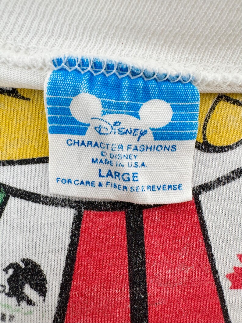 Może przedstawiać: Zbliżenie białego kołnierzyka koszulki z niebiesko-białą metką Disney Character Fashions. Na metce widnieje napis "LARGE" i "MADE IN U.S.A.". Koszulka ma czerwono-biały wz&oacute;r.