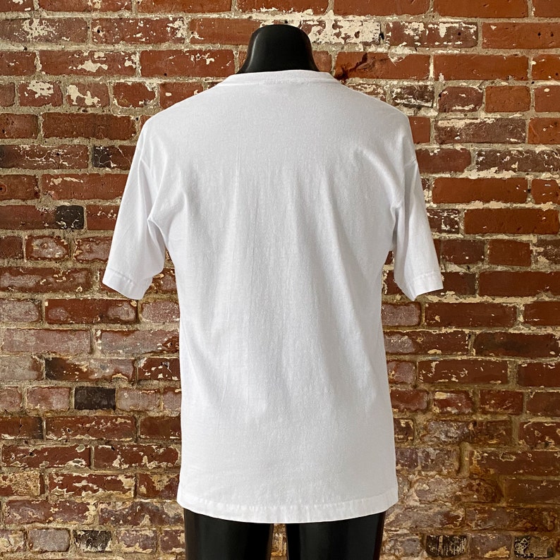 May include: A plain white t-shirt with short sleeves. The shirt is on a mannequin against a brick wall background.