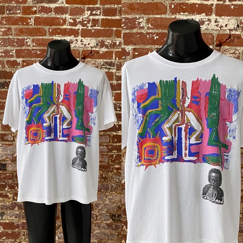May include: White t-shirt with a colorful abstract design featuring a sun and two figures. The design is in shades of pink, blue, green, yellow, and red. The shirt also features a small black and white image of a person with the word "miles" printed below.