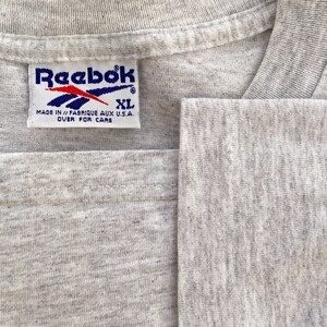 May include: A gray Reebok t-shirt tag with white text that reads "Reebok" and "Made in // Fabrique aux U.S.A. Over for care". The tag also has a blue Reebok logo and the size "XL" in blue.