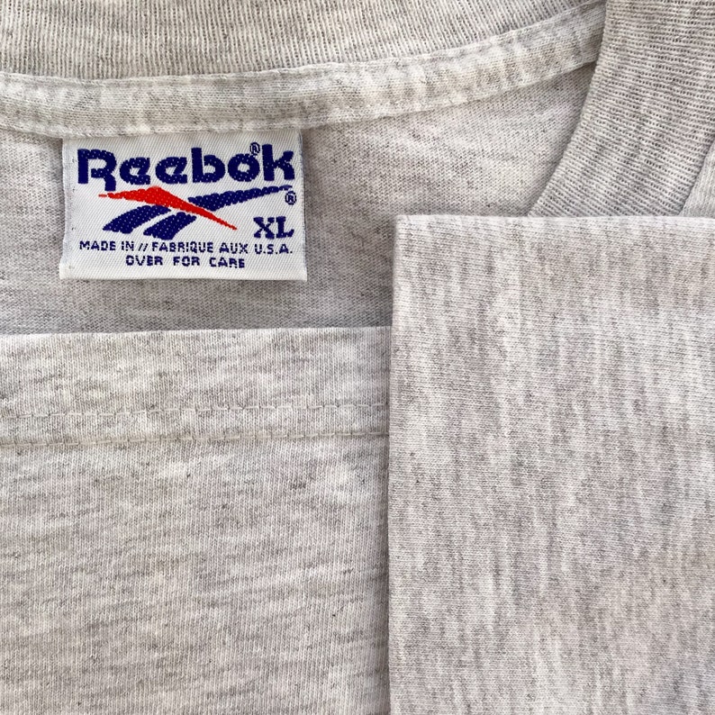 May include: A gray Reebok t-shirt tag with white text that reads "Reebok" and "Made in // Fabrique aux U.S.A. Over for care". The tag also has a blue Reebok logo and the size "XL" in blue.