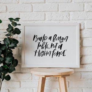 May include: White framed print with black calligraphy text on a white background. The text reads "Build a longer table, not a higher fence". The print is hanging on a white brick wall with a small wooden stool in front of it.