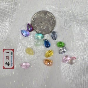 May include: Assortment of colorful glass beads in various shapes and sizes, including teardrop, oval, and round. The beads are scattered on a white surface with a US quarter for scale.