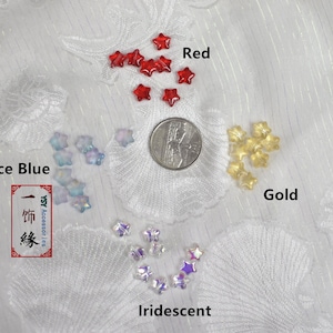 May include: Assortment of star-shaped glass beads in various colors, including red, ice blue, gold, and iridescent. The beads are arranged in four groups on a white fabric background. A US quarter is included for size reference.