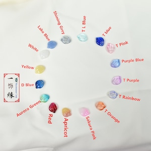 May include: A variety of small, colorful shell-shaped charms in different colors, including white, yellow, blue, green, red, apricot, pink, orange, and purple. The charms are arranged in a circle on a white fabric background. The text "YSY Accessories 一饰緣" is visible in the lower left corner of the image.