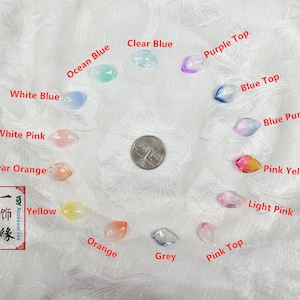 May include: Assortment of 14 clear glass beads in various colors, including white, pink, orange, yellow, blue, purple, and grey. The beads are shaped like teardrops and have a smooth, glossy finish. The beads are arranged in a circle on a white surface. A US quarter is included for scale.