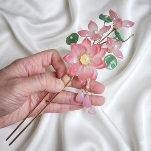 Peut inclure: Une épingle à cheveux décorative avec un motif de fleur de lotus rose, des feuilles vertes et des perles. L'accessoire pour cheveux a une épingle dorée et des breloques roses en forme de goutte. Le motif floral est sur fond blanc.