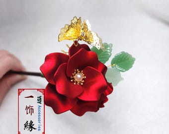 May include: A vibrant red flower hair accessory with a gold butterfly accent and green leaves. The flower has a pearl center and is attached to a dark stem. The accessory is likely for a special occasion.