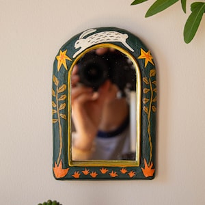 May include: An arched wall mirror with a dark green frame featuring a hand-painted white rabbit at the top, yellow stars on the sides, and orange floral motifs along the bottom and vertical stems. The inner edge of the frame is painted gold.