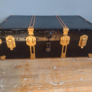 Puede incluir: Un ba&uacute;l vintage negro con detalles dorados y herrajes de lat&oacute;n. El ba&uacute;l tiene dos pestillos a cada lado y una cerradura en el centro.