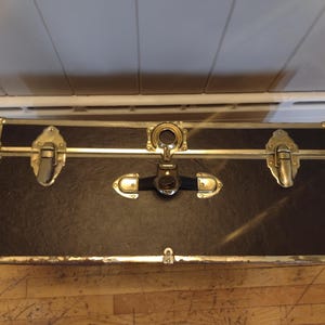 May include: A rectangular black trunk with gold-colored metal accents and hardware. The trunk has a textured surface and is secured with clasps and a central lock. The corners are reinforced with metal.