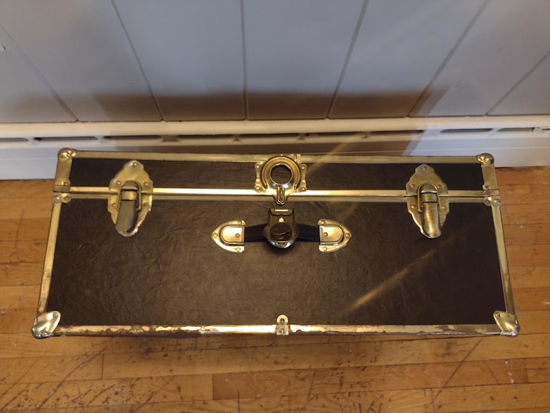 May include: A rectangular black trunk with gold-colored metal accents and hardware. The trunk has a textured surface and is secured with clasps and a central lock. The corners are reinforced with metal.