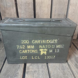 May include: An olive green metal ammunition box with a hinged lid and a handle. The box has yellow stenciled text that reads "200 CARTRIDGES 7.62 MM NATO M82 CARTONS M 13 LOT LCL 13017".