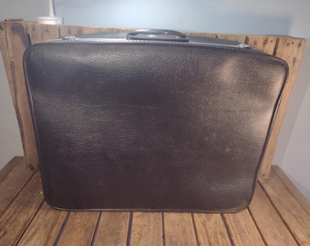 May include: A vintage black suitcase with a textured surface. The suitcase has a silver handle and trim. It is sitting on a wooden surface.