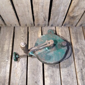 May include: A vintage green metal cheese grater with a wooden handle and a crank. The grater is sitting on a wooden surface.
