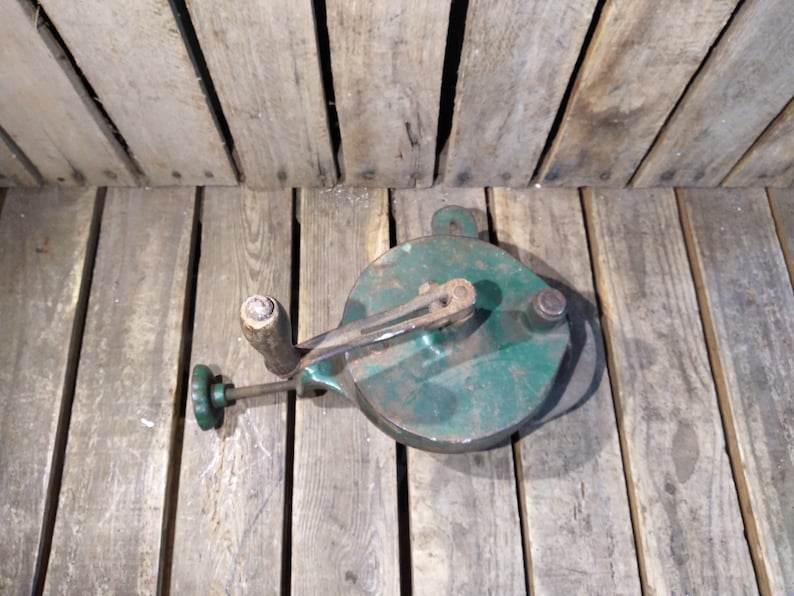 May include: A vintage green metal cheese grater with a wooden handle and a crank. The grater is sitting on a wooden surface.