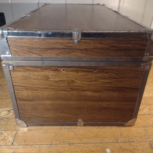 May include: A brown wooden storage trunk with metal accents and a hinged lid. The trunk has two metal clasps on the front and two metal handles on the sides.