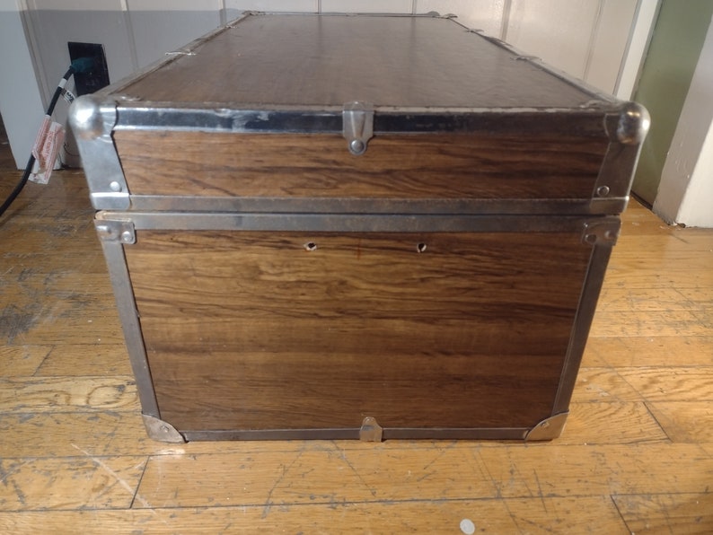 May include: A brown wooden storage trunk with metal accents and a hinged lid. The trunk has two metal clasps on the front and two metal handles on the sides.