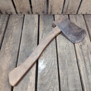 May include: A vintage axe with a wooden handle and a rusty metal head. The axe is lying on a wooden surface, showcasing its aged appearance and classic design. The axe's head is approximately 6 inches long.