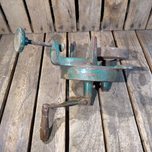 May include: A vintage green metal hand crank tool with a wooden handle and a metal clamp. The tool is on a wooden surface.