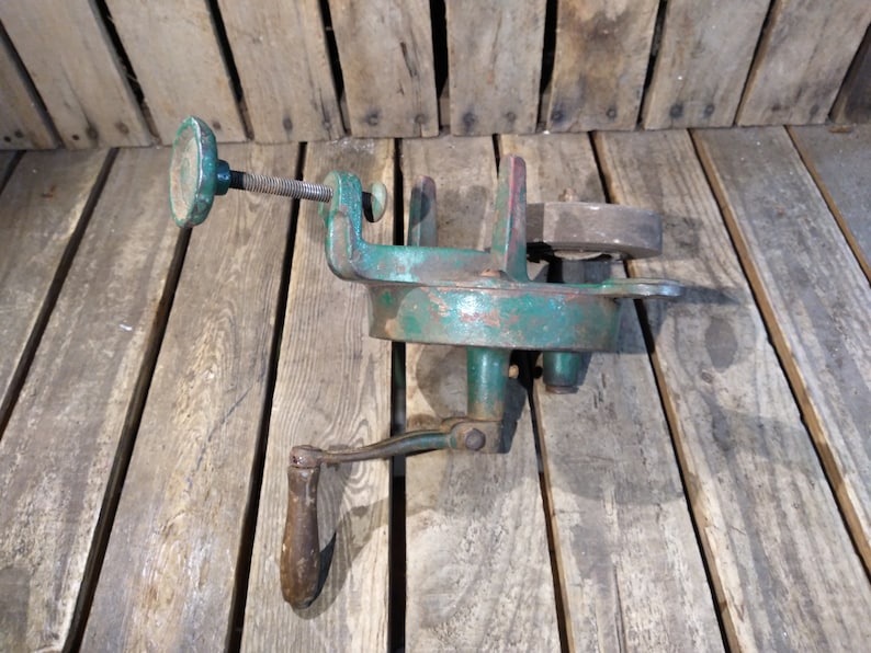 May include: A vintage green metal hand crank tool with a wooden handle and a metal clamp. The tool is on a wooden surface.