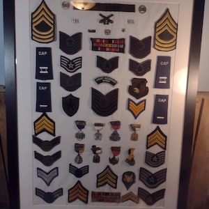 May include: Framed collection of military insignia, including chevrons, ribbons, and badges, displayed on a white background. The insignia feature various colors and designs, showcasing ranks and achievements. The frame is black.