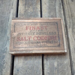 May include: A vintage wooden box with the text "FINAST BRAND ENTIRELY BONELESS SALT CODFISH" in red and black lettering. The box is labeled "ONE POUND NET WEIGHT" and "PACKED IN CANADA". The box is on a wooden surface.