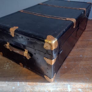 Puede incluir: Ba&uacute;l vintage negro con herrajes de lat&oacute;n y asa de madera. El ba&uacute;l tiene un aspecto desgastado y envejecido y est&aacute; hecho de material resistente.
