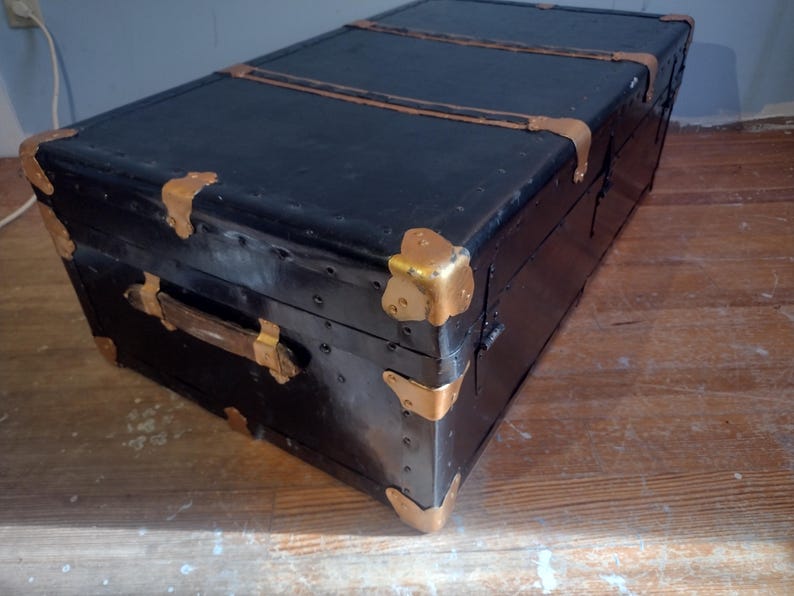 Puede incluir: Ba&uacute;l vintage negro con herrajes de lat&oacute;n y asa de madera. El ba&uacute;l tiene un aspecto desgastado y envejecido y est&aacute; hecho de material resistente.
