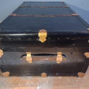 Puede incluir: Un ba&uacute;l de viaje vintage negro con detalles dorados y un asa de cuero. El ba&uacute;l tiene un aspecto desgastado y envejecido y est&aacute; hecho de madera.