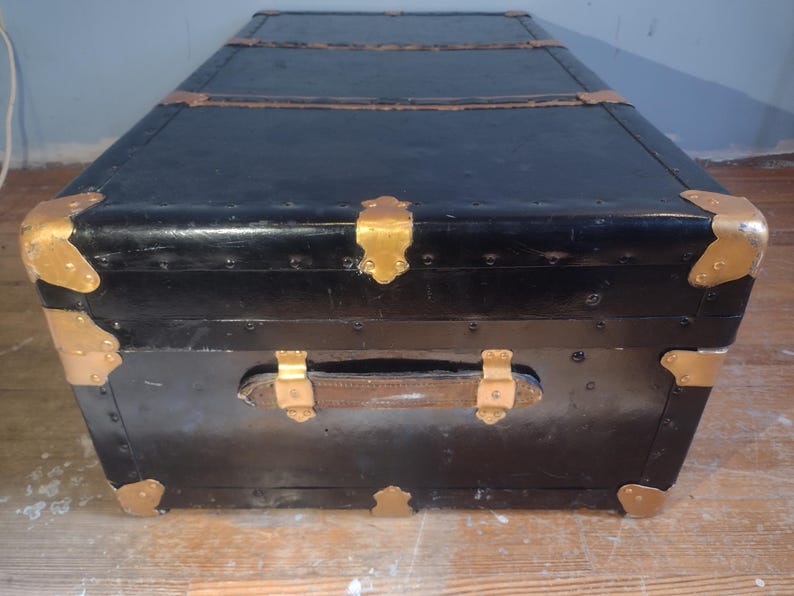 Puede incluir: Un ba&uacute;l de viaje vintage negro con detalles dorados y un asa de cuero. El ba&uacute;l tiene un aspecto desgastado y envejecido y est&aacute; hecho de madera.
