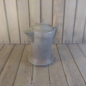 Peut inclure: Une cruche en argent avec un couvercle et une poignée. La cruche est posée sur une surface en bois.