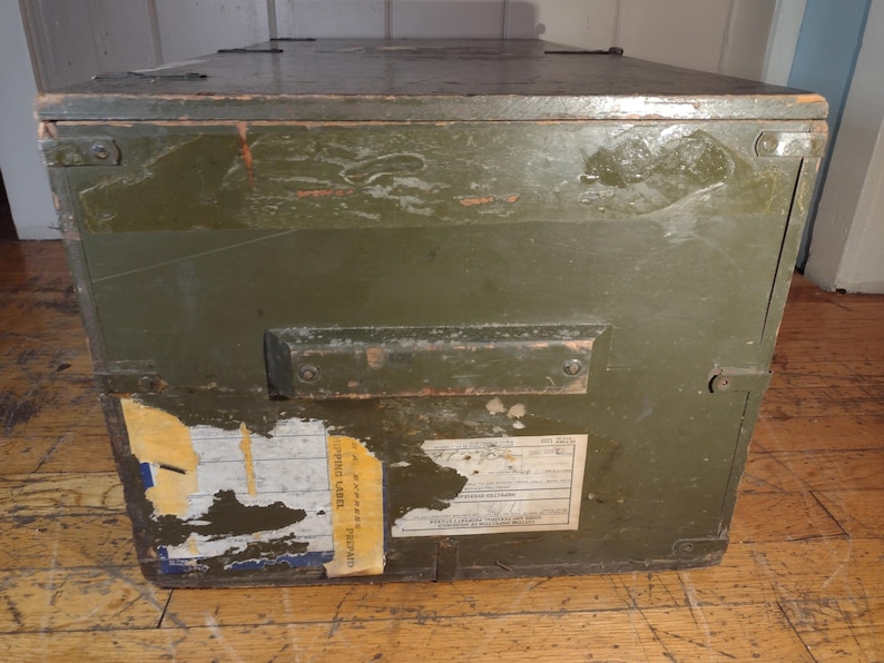 May include: A vintage green wooden storage box with a metal handle and a yellow shipping label. The label has the text "SHIPPING LABEL" and "NECESSARY" printed on it.
