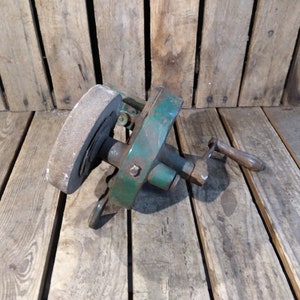 May include: A vintage green metal hand-crank sharpening tool with a round gray stone wheel. The tool has a wooden handle and is resting on a wooden surface.