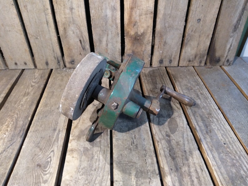May include: A vintage green metal hand-crank sharpening tool with a round gray stone wheel. The tool has a wooden handle and is resting on a wooden surface.