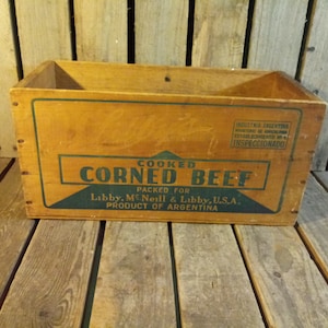 Könnte beinhalten: Eine Holzkiste mit einem grün-weißen Etikett, auf dem "Cooked Corned Beef Packed for Libby, McNeill & Libby, U.S.A. Product of Argentina" steht. Die Kiste steht auf einer Holzoberfläche.