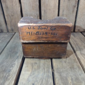 May include: A vintage wooden tool box with the text "U.S. Tool Co." and "MT-8195-101" in black lettering. The box is rectangular with a notched top and is made of aged, weathered wood. The background is a wooden surface.