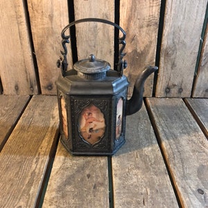 May include: A black metal teapot with a decorative handle and a lid. The teapot has a square body with a floral design and a painted scene on each side.