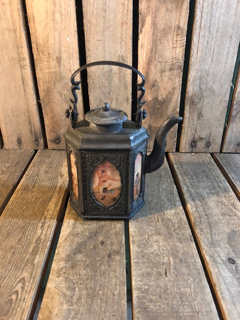 May include: A black metal teapot with a decorative handle and a lid. The teapot has a square body with a floral design and a painted scene on each side.