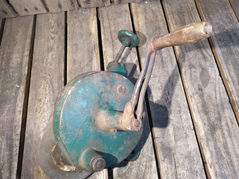 May include: A vintage green metal hand crank grinder with a wooden handle. The grinder has a round base and a metal crank with a green knob.