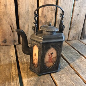 May include: A black metal teapot with a hinged lid and a decorative handle. The teapot has a square body with four panels, each featuring a painted scene of a person in traditional clothing.