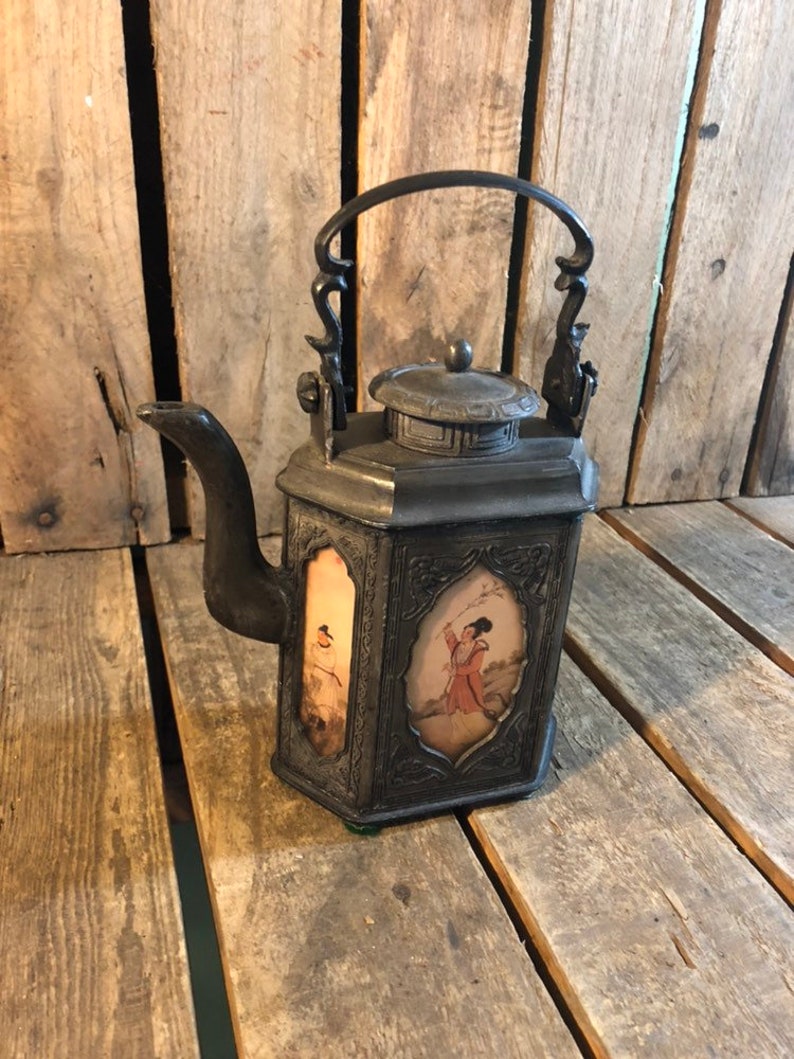 May include: A black metal teapot with a hinged lid and a decorative handle. The teapot has a square body with four panels, each featuring a painted scene of a person in traditional clothing.