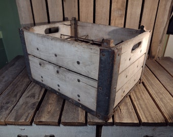 Op de afbeelding: Een vintage houten kist met metalen banden. De kist is verweerd en heeft een witgekalkte afwerking. Hij heeft een metalen handvat aan de zijkant en staat op een houten oppervlak.