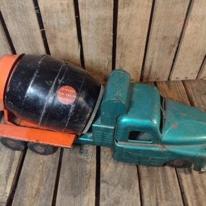 May include: Vintage toy cement mixer truck in teal and orange. The truck features a black rotating drum with a red and white sticker. The cab is teal, and the mixer's frame is orange. The toy is made of metal.