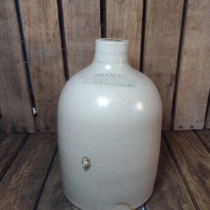 May include: A vintage stoneware jug with a rounded body and a narrow neck. The jug is a light beige color with a few imperfections and markings. The text "FORTMAN BROS. FORT LUDLOW, W.VA." is visible on the jug.